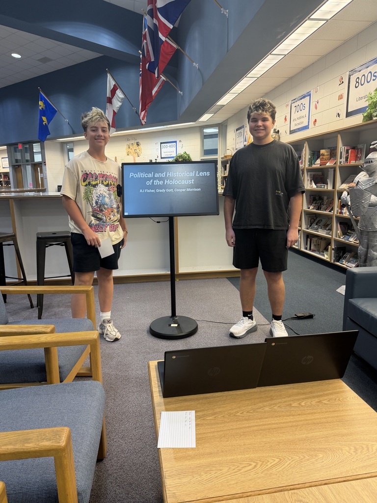 Two people stand next to a monitor in a room with flags, a table, and chairs.