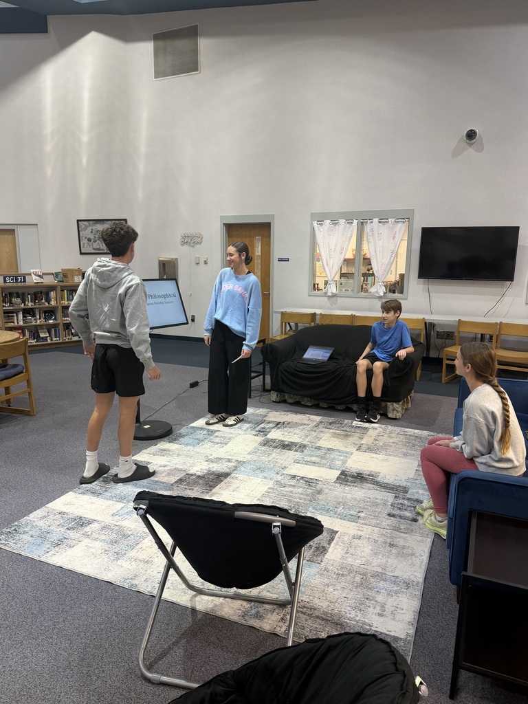 Four people in a room with a rug. One person stands with a microphone. Two sit on a couch.