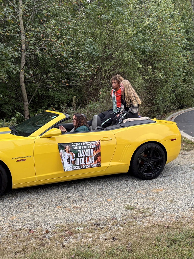 Two people ride in a yellow convertible car with a banner. Trees surround the scene.