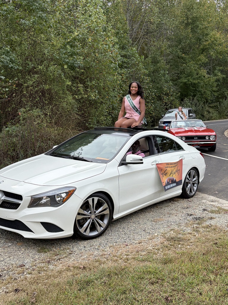 A white car is parked on a side road with a woman in a sash sitting on the hood. Trees and grass are in the background.