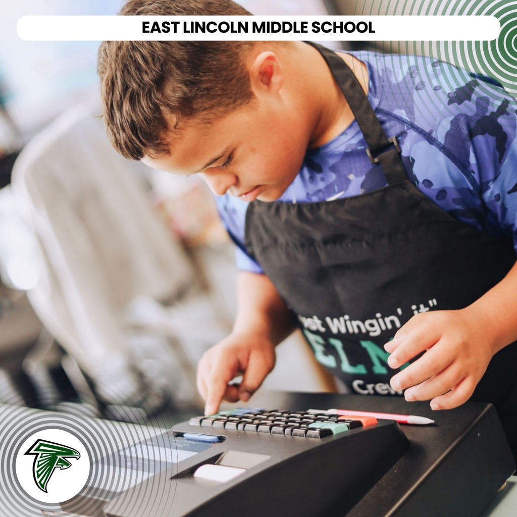 A young boy in a blue shirt and black apron types on a cash register in a classroom.