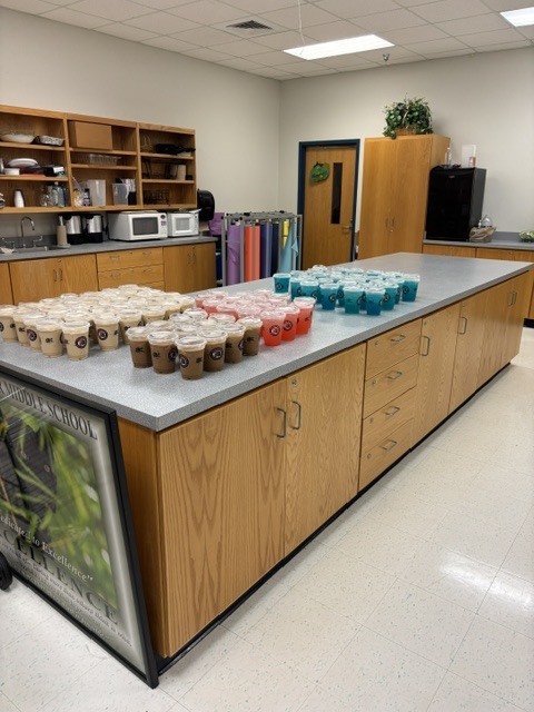 A kitchen area with a large counter holding numerous cups of beverages. Wooden cabinets and shelves line the walls.