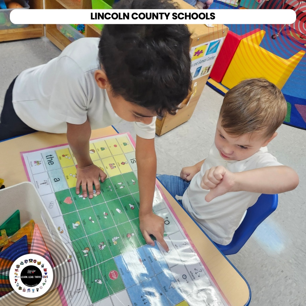 Two young children sit at a table, one pointing at a game board while the other looks on.