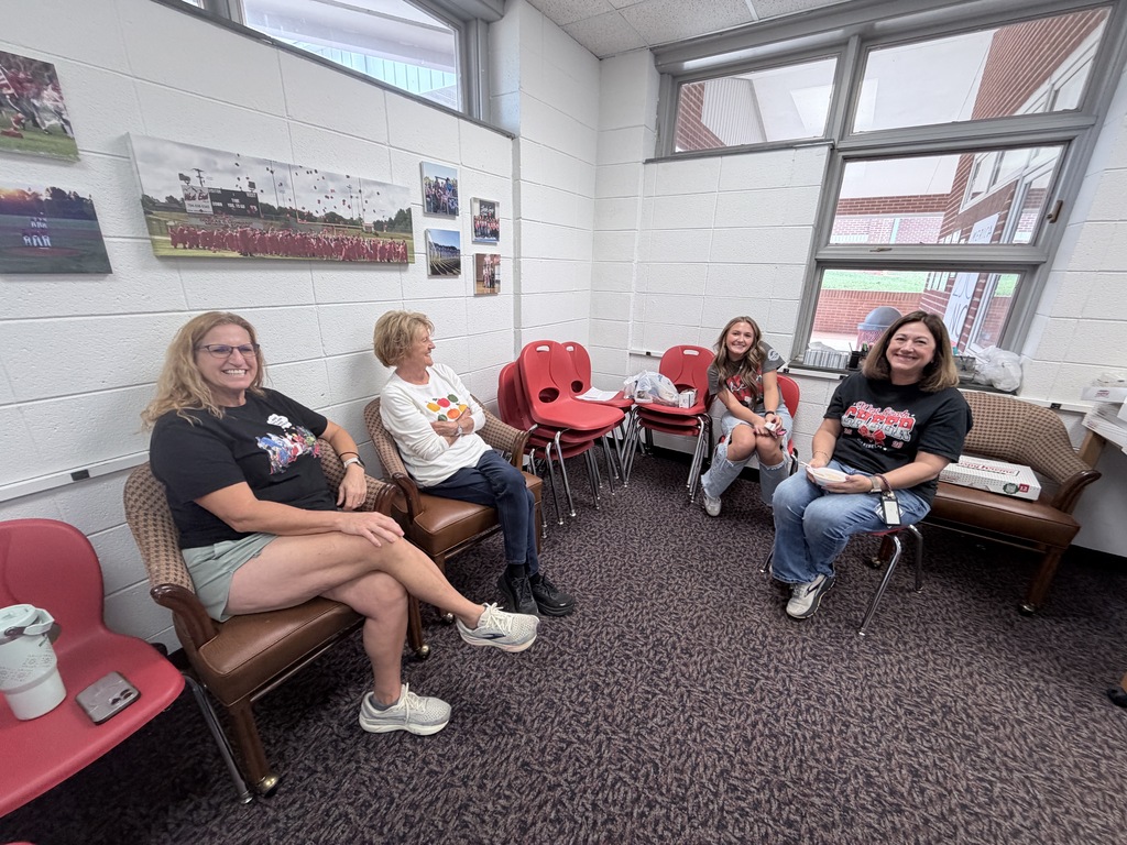 A group of women sit on chairs in a room with carpeting, chairs, and a couch. Behind them, a white wall has pictures.