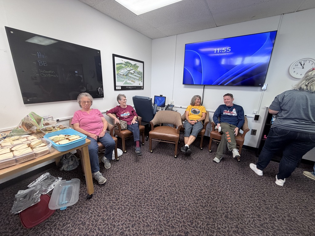 Six people in a room with chairs. A woman stands near a table with food. A clock is on the wall.