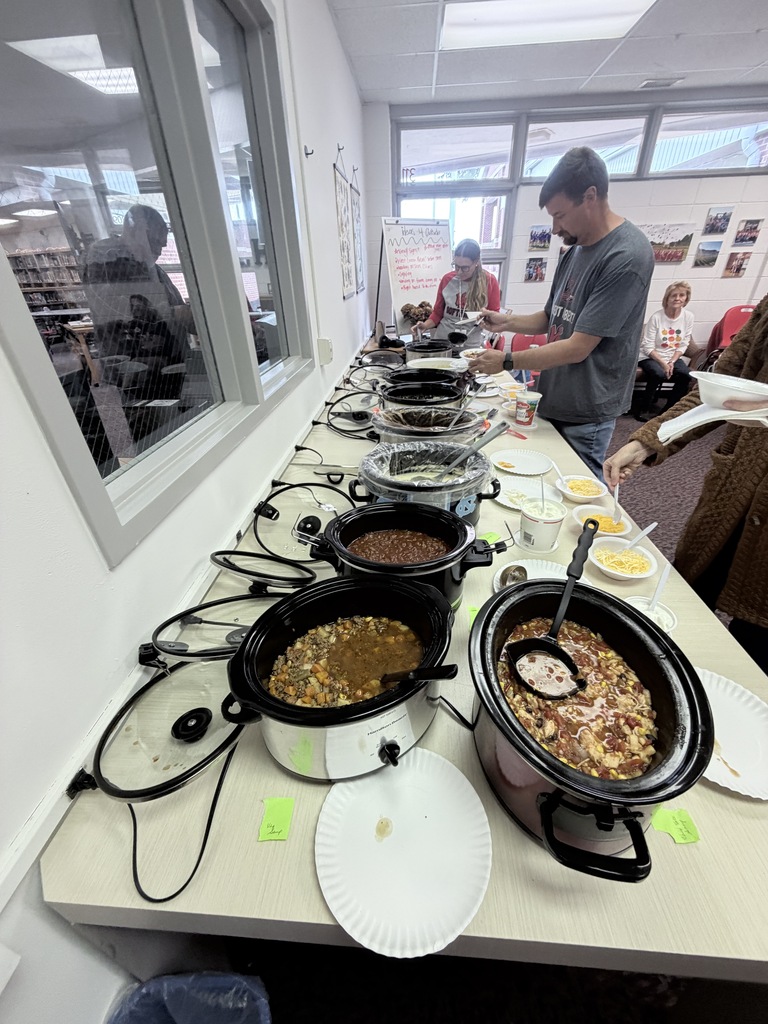 A room with a long table, several slow cookers, plates, and people standing or seated.