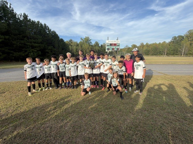 Youth soccer team poses on a field, wearing matching white uniforms and sneakers, with a scoreboard visible in the background.