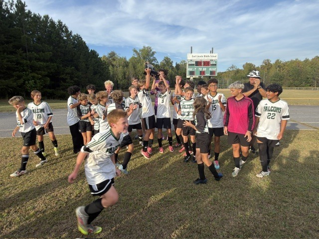Group of kids in white and black soccer uniforms celebrating with a trophy on a grassy field.