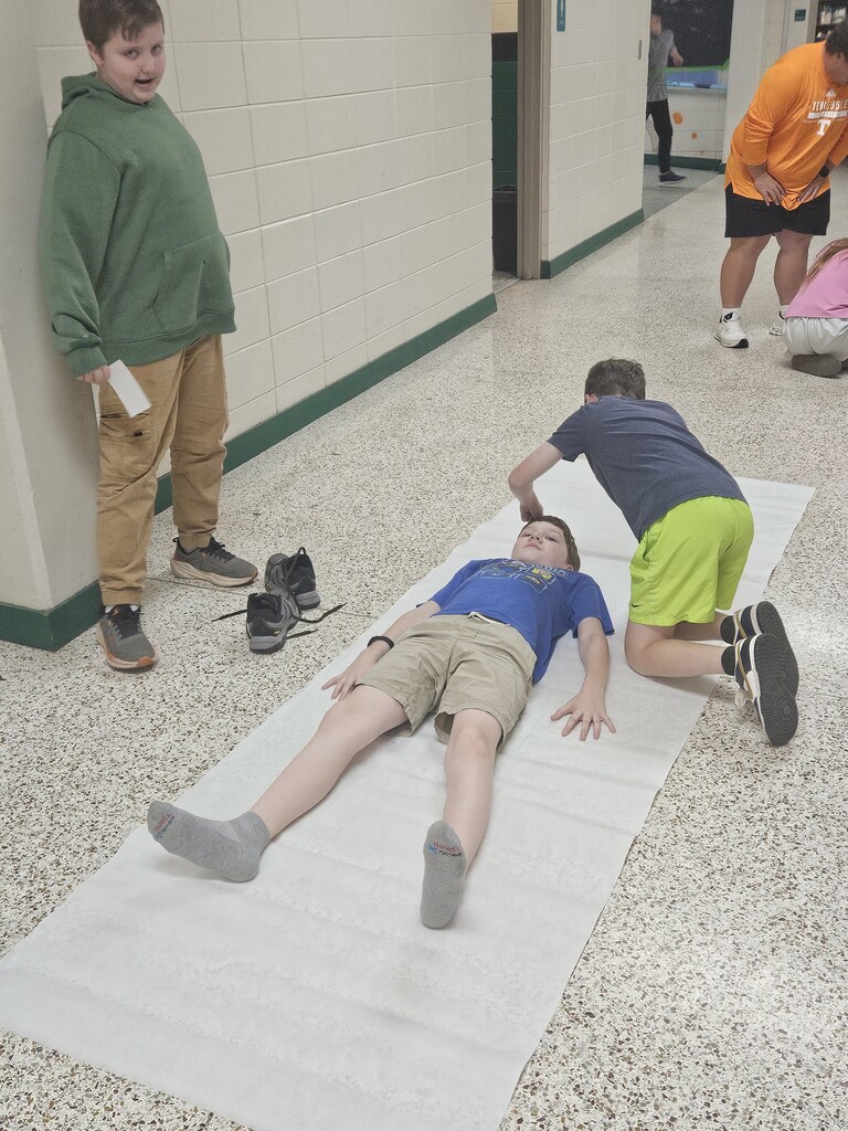 Three boys in a hallway. One boy lies on a white mat, another kneels over him, and the third stands near a wall.