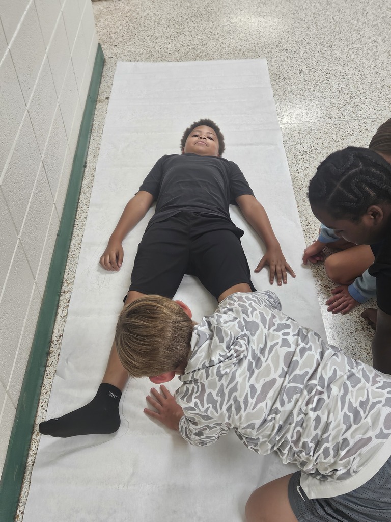 A person lies on a white sheet on a floor. Two people kneel nearby, one checking the person's leg.