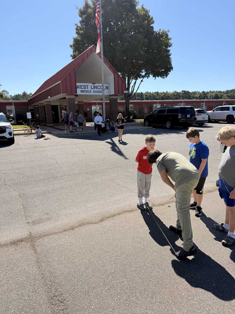 Several children stand outside a building, one bends down while another watches. Cars park behind.
