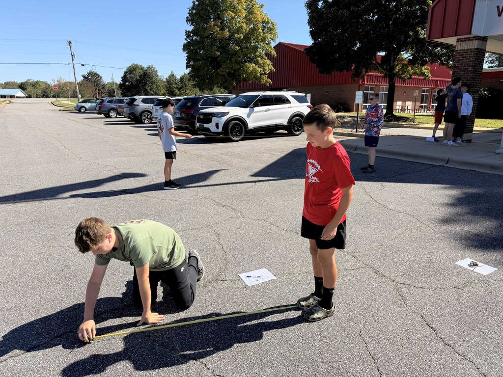 A boy in a green shirt measures on the pavement with a tape measure. A boy in red stands in front of him.