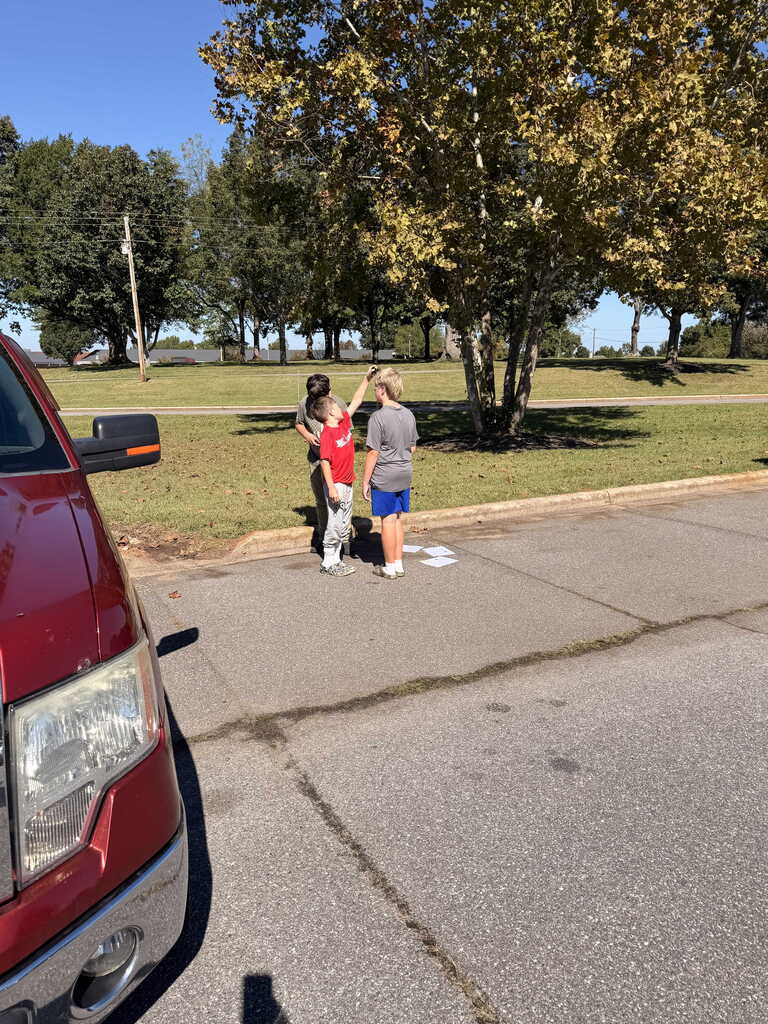 Two people stand by a tree in a park, one with a red shirt. A red car is parked nearby.