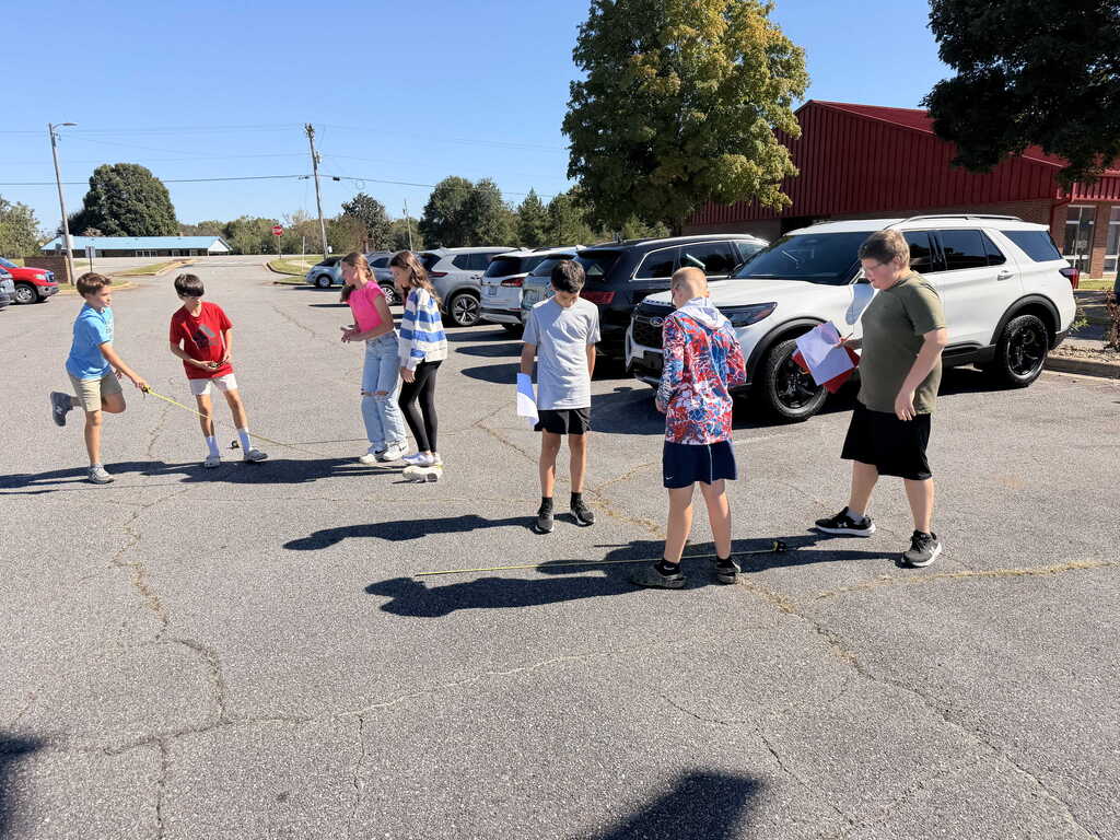 Several people in a parking lot, some walking and others standing. Vehicles parked and a red building in the background.