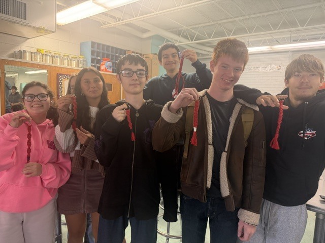 A group of six high school students standing in a classroom holding up handmade red macramé keychains.