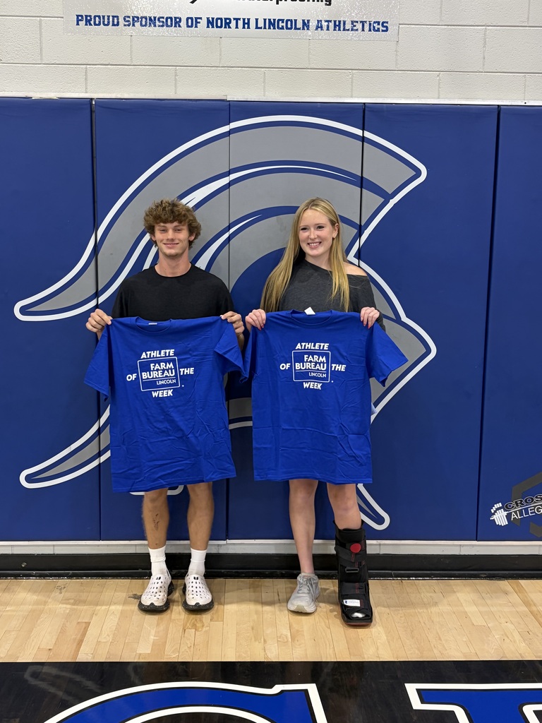 Two young people in a gymnasium, holding up blue shirts, one wearing a black shirt and the other a gray shirt.