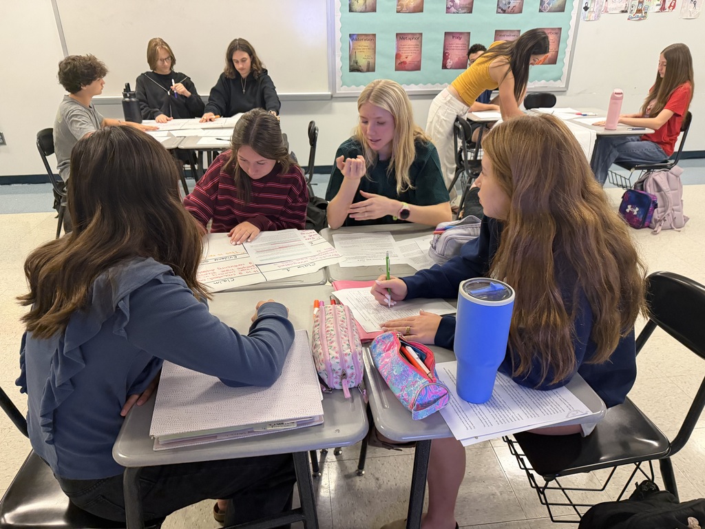 Students in a classroom with desks, books, and supplies, seated around tables, engaged in academic activities.