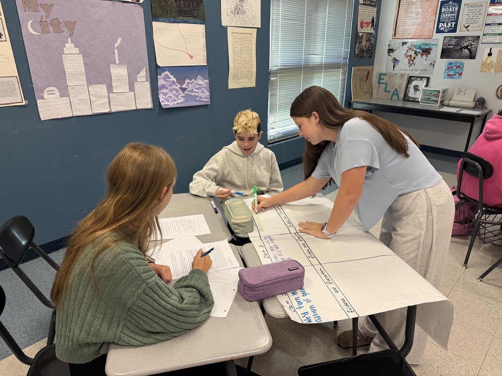 Three students work on a large piece of paper on a table, one seated and two standing.