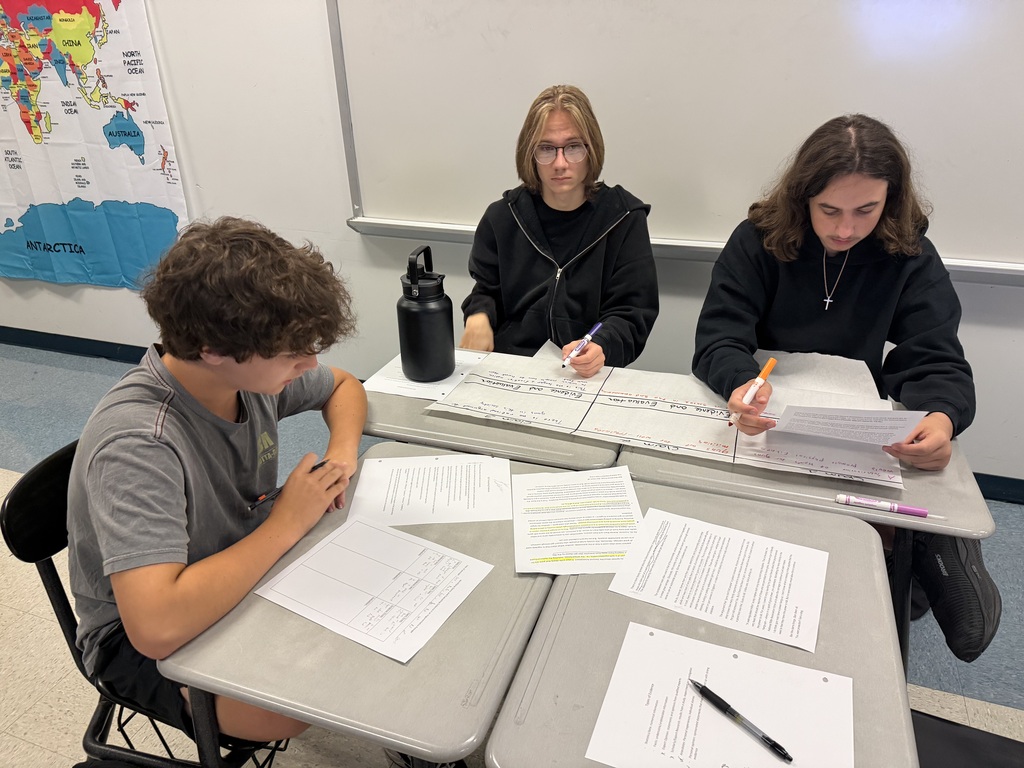 Three students are seated at desks with papers and pens, one wearing glasses and the others in black jackets.
