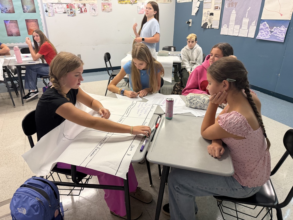 Students sit at desks and work on a project in a classroom, with one student looking at a large sheet of paper.