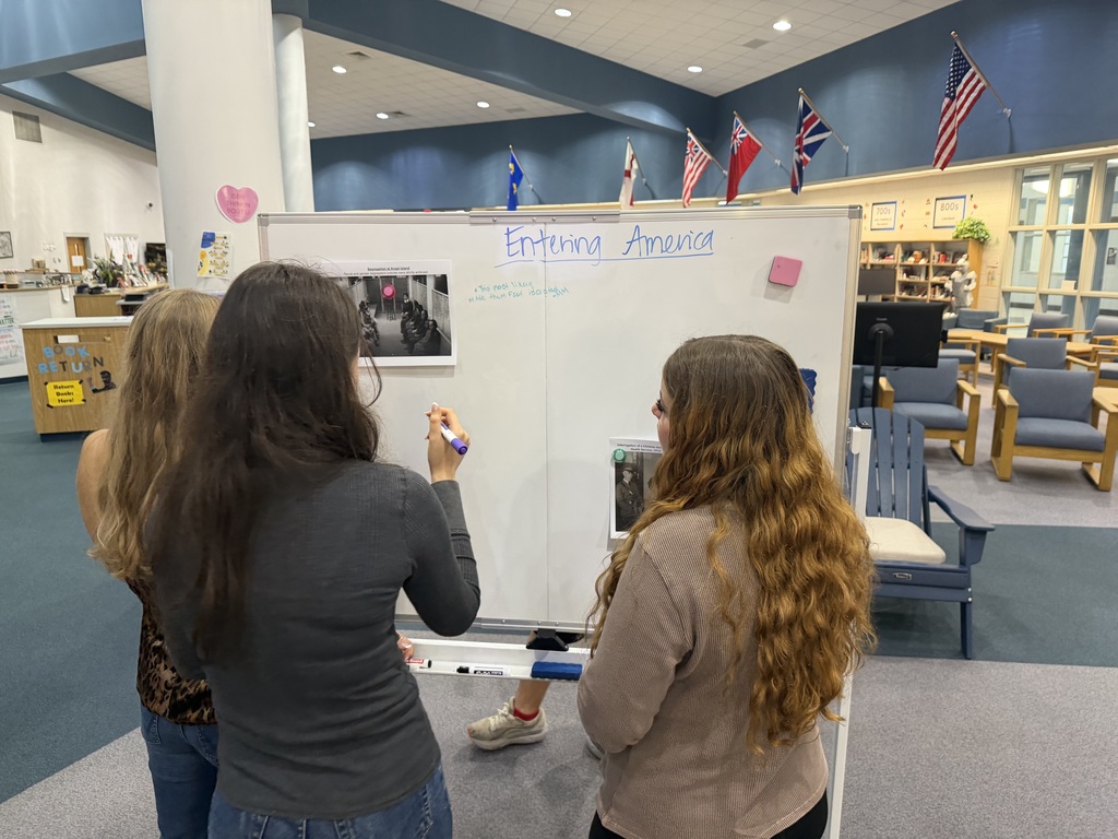 Three people write on a whiteboard in a library. Flags and chairs are in the background.