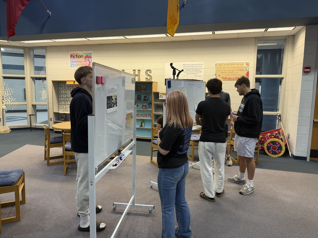 Students gather in a classroom, looking at a whiteboard, with bookshelves and tables in the background.