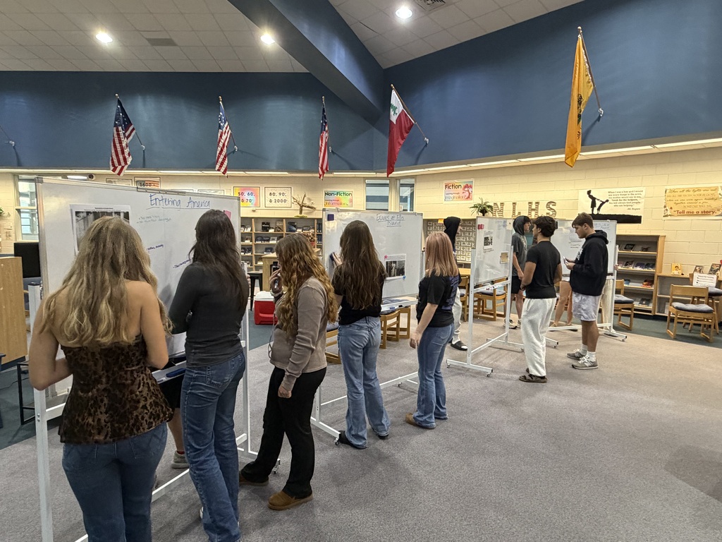 A group of people stand in a room with white boards, two flags, and bookshelves.
