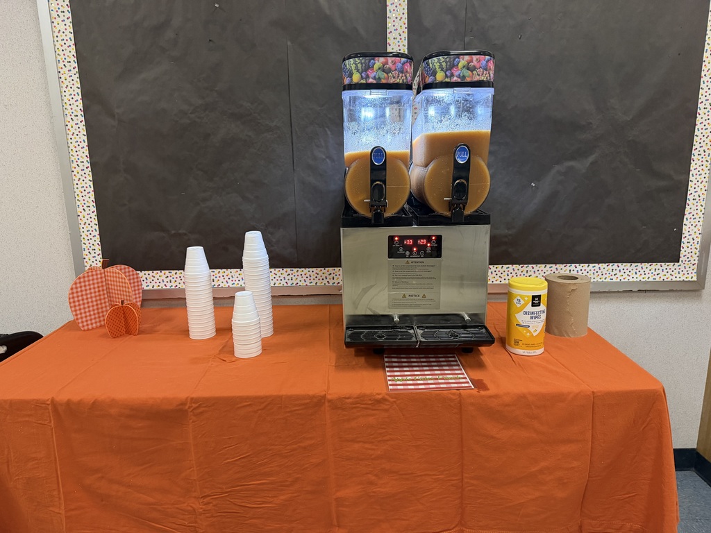 An orange tablecloth covers a table with a coffee machine and cups. A blackboard hangs on the wall behind the machine.
