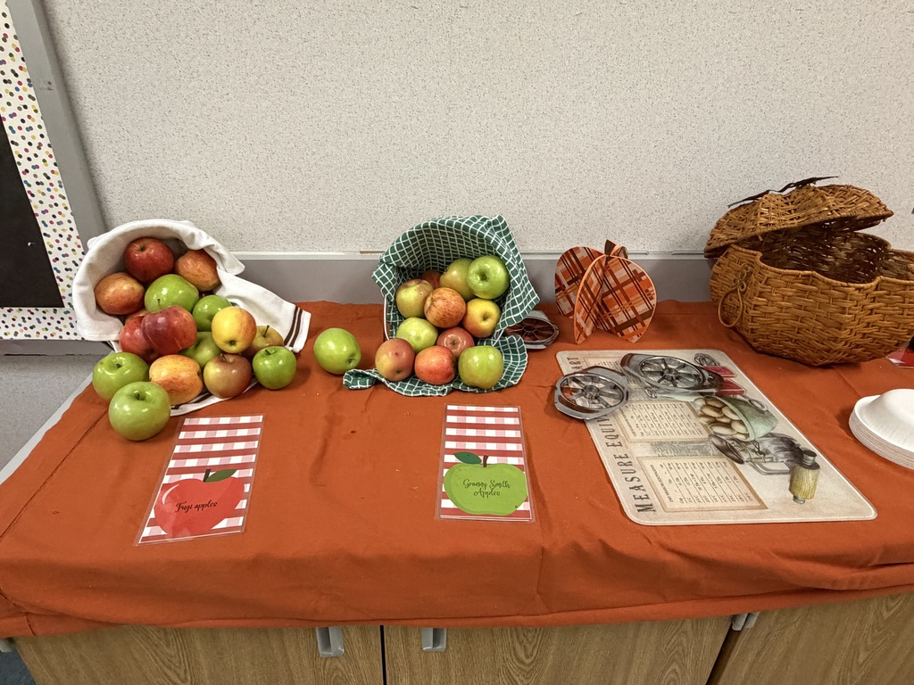 A table with an orange tablecloth has several apples in bags, a basket, and other items.