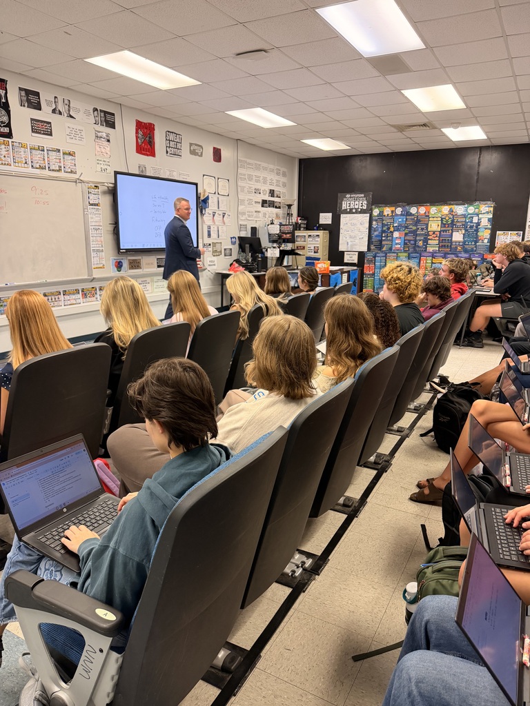 A teacher stands before a screen, teaching a seated classroom audience. Each student is using a laptop.