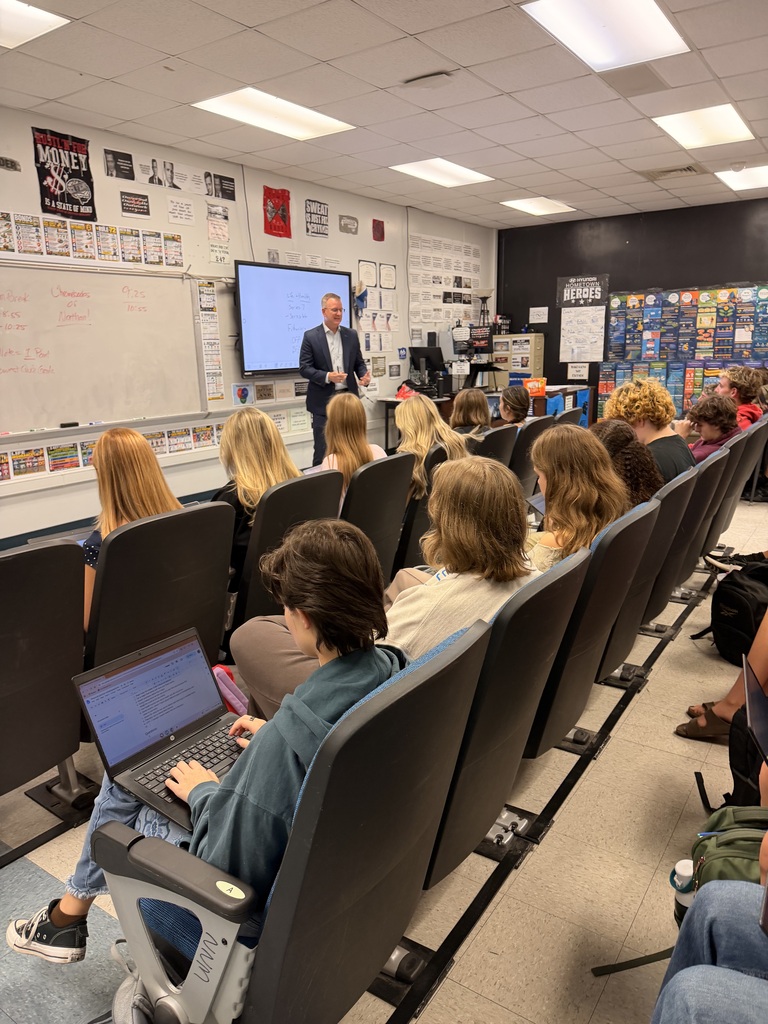 A classroom with a person giving a presentation on a screen. Many students are seated in chairs, attentively watching.