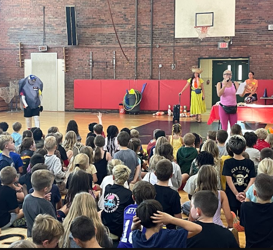 A group of students gathered in a gym for an event. Speakers on stands and a basketball hoop are visible.