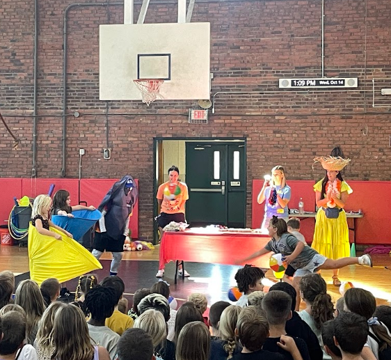 Audience watching performers in a gymnasium. One performer has a hat and a yellow skirt.