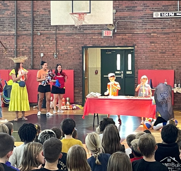 Several people in a gym performing a show with a red table in front of an audience.