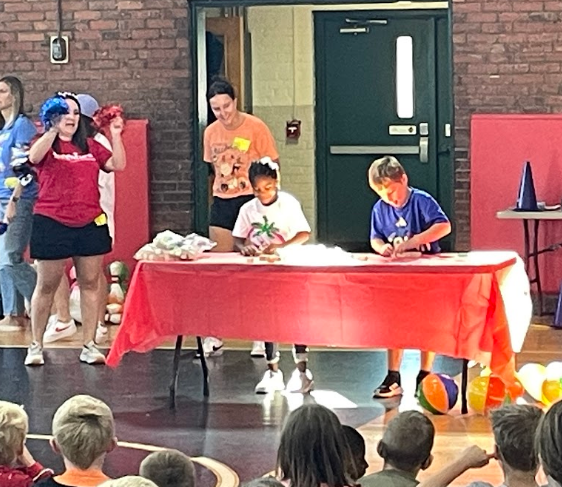A table with a red tablecloth has two children building towers. A woman stands before them.