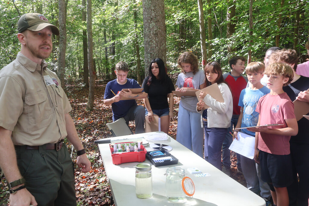 A group of students listen to a man in a forest. A desk holds various items, and students hold clipboards.