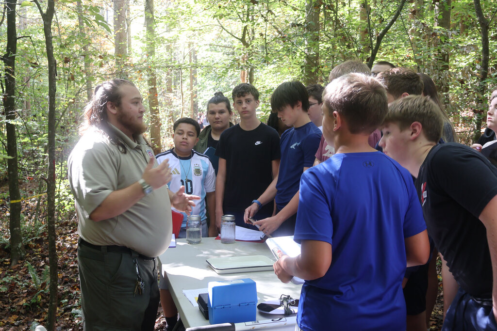Group of students in a forest setting, with one adult leading a discussion at a table.