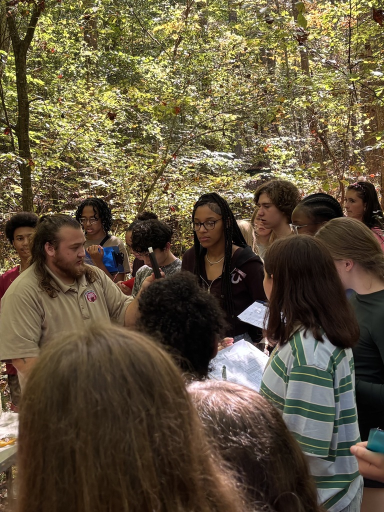 Group of individuals in a forest. One man stands at the center, speaking. Various people look in his direction.