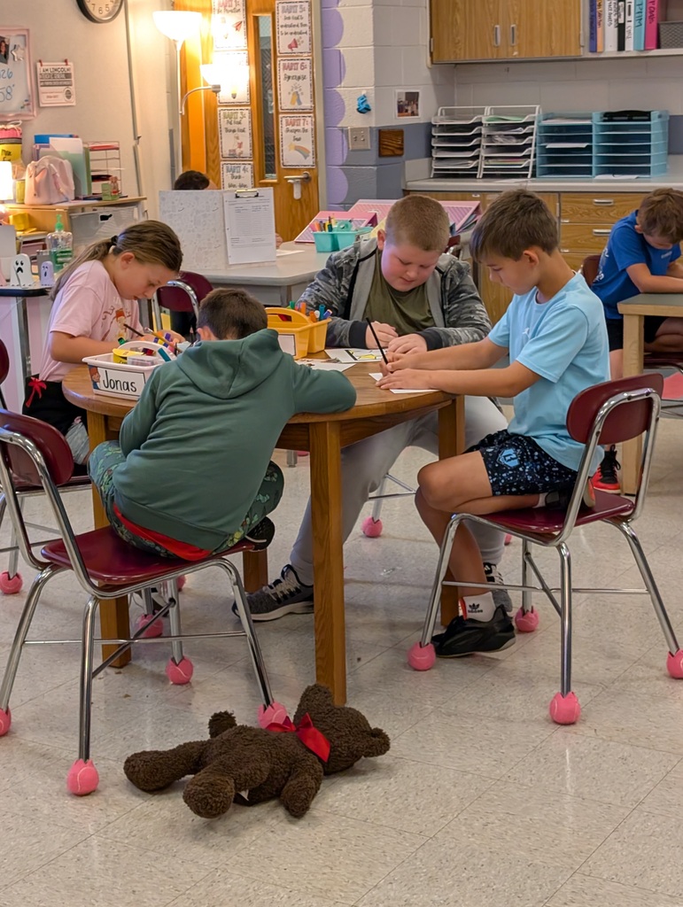 Four children in a classroom, seated around a table, engaged in coloring and writing, with a stuffed bear on the floor.
