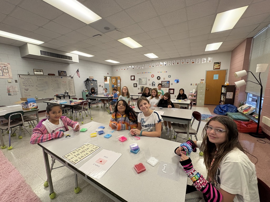Four children, one with glasses, sit at a table in a classroom. They have various items and papers on the table.