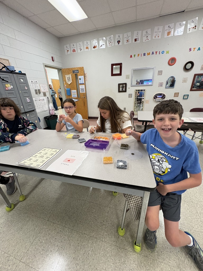 Four children seated around a table in a classroom, engaged in an activity. A boy in a blue shirt is leaning forward, smiling.