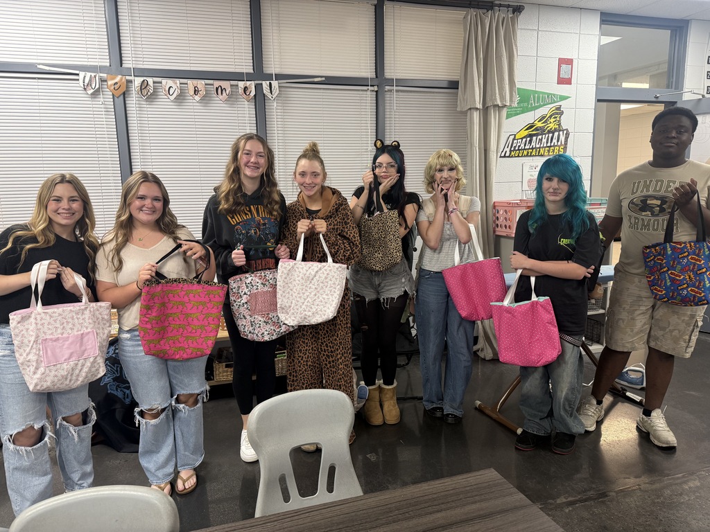 Nine people are posing for a photo while holding colorful bags. A white chair is behind them.