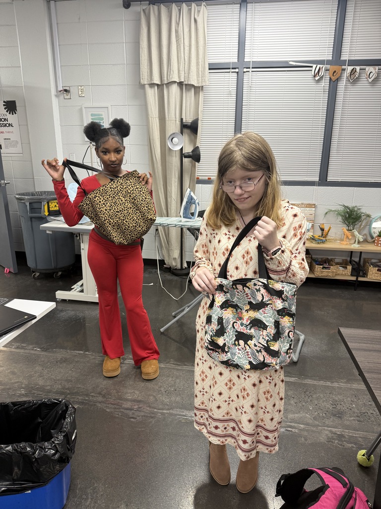 Two women stand in a room with bags. One holds a large bag, the other a smaller one. Behind them, a table has an iron, and storage boxes are on a shelf.