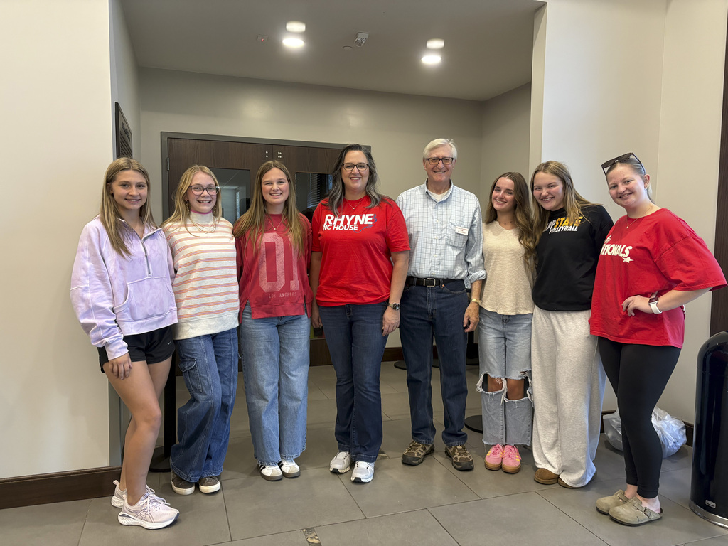 Group of eight people standing side-by-side indoors. From left to right: a young woman in a light purple quarter-zip sweatshirt and black shorts; a young woman in a striped long-sleeve shirt and blue cargo jeans; a young woman in a red long-sleeve top and blue jeans; a woman in a red "RHYNE NC HOUSE" t-shirt and jeans; an older man in a light blue button-down shirt, dark pants, and glasses; a young woman in a beige top and ripped jeans; a young woman in a black "App State Volleyball" shirt and light gray sweatpants; and a young woman in a red t-shirt with "Nationals" text and black leggings.