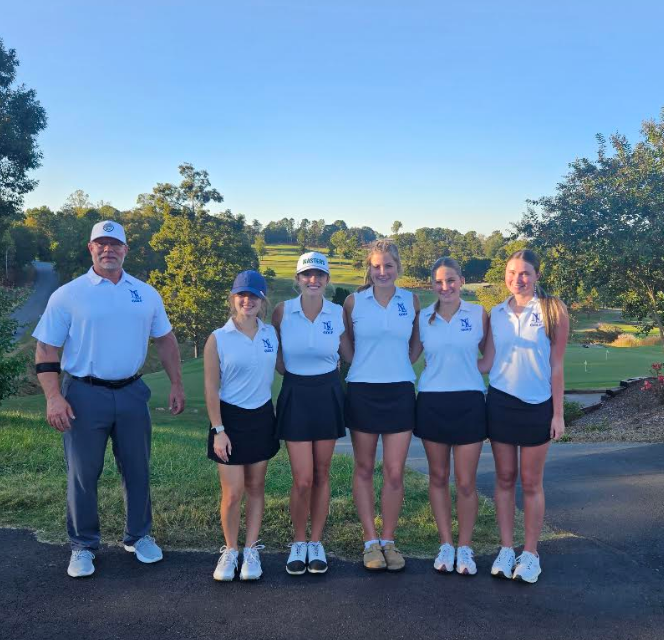 A male coach and five female athletes stand in a row on a road, all wearing matching white shirts and black skirts.