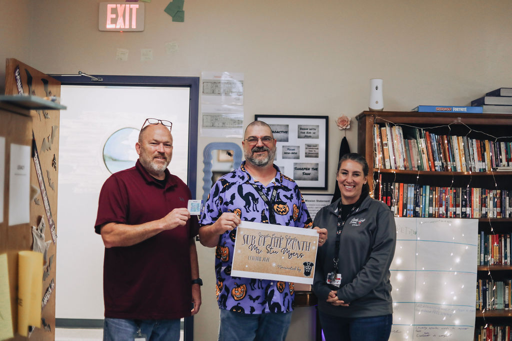 Three people in a room with bookshelves, holding a sign that reads "Volunteer of the Month."