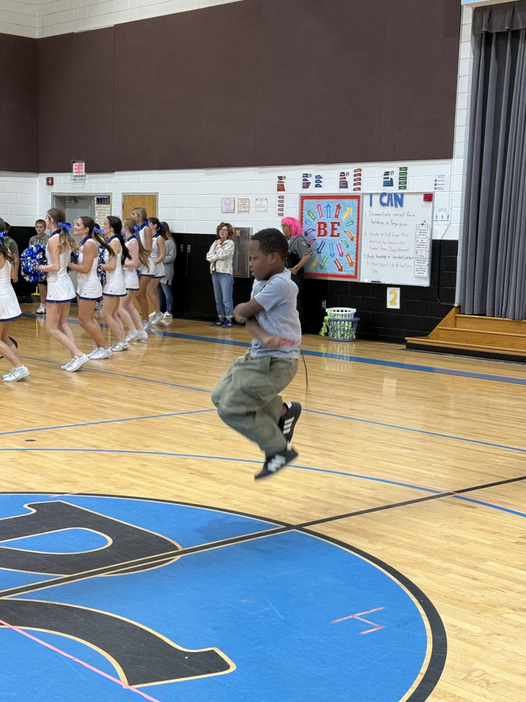 A boy in the air jumping rope while a line of cheerleaders in white uniforms stands behind him.