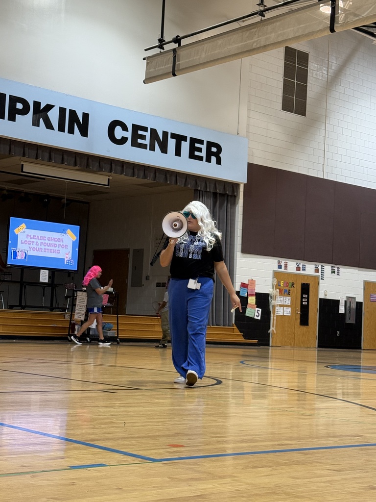 Person with white wig and megaphone walking on a basketball court, with building reading "Pipkin Center" in background.