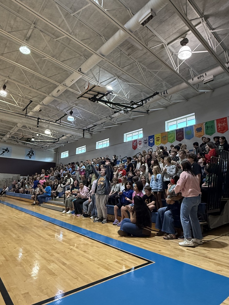 A gymnasium filled with many people seated on bleachers, a wooden floor, and blue lines on the court.
