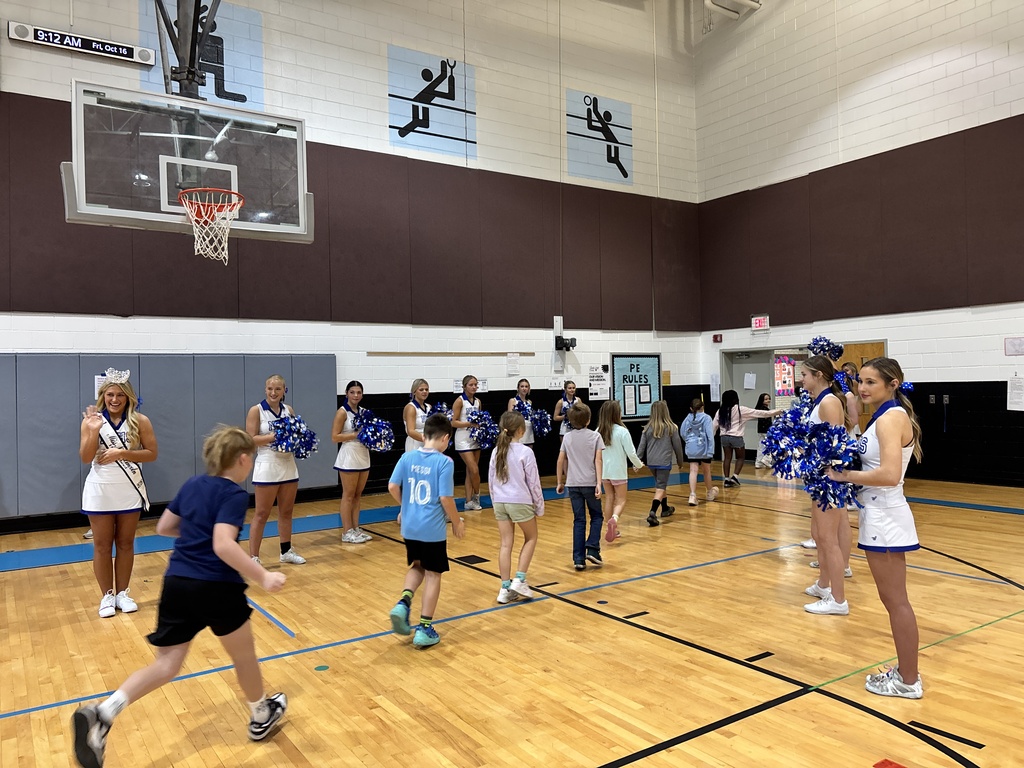 A group of children in uniforms play a game on a basketball court, with cheerleaders holding pom-poms.